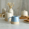 Habulous small stoneware mug cornflower blue in front of large white mug and table setting of white stoneware ceramics