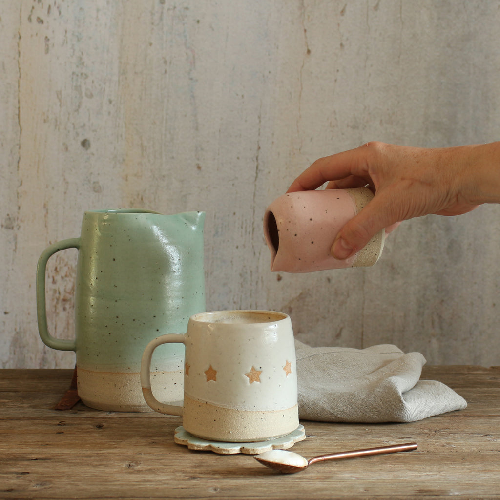 Star mug with hand pouring pink milk jug to make coffee with a green pitcher jug in the background