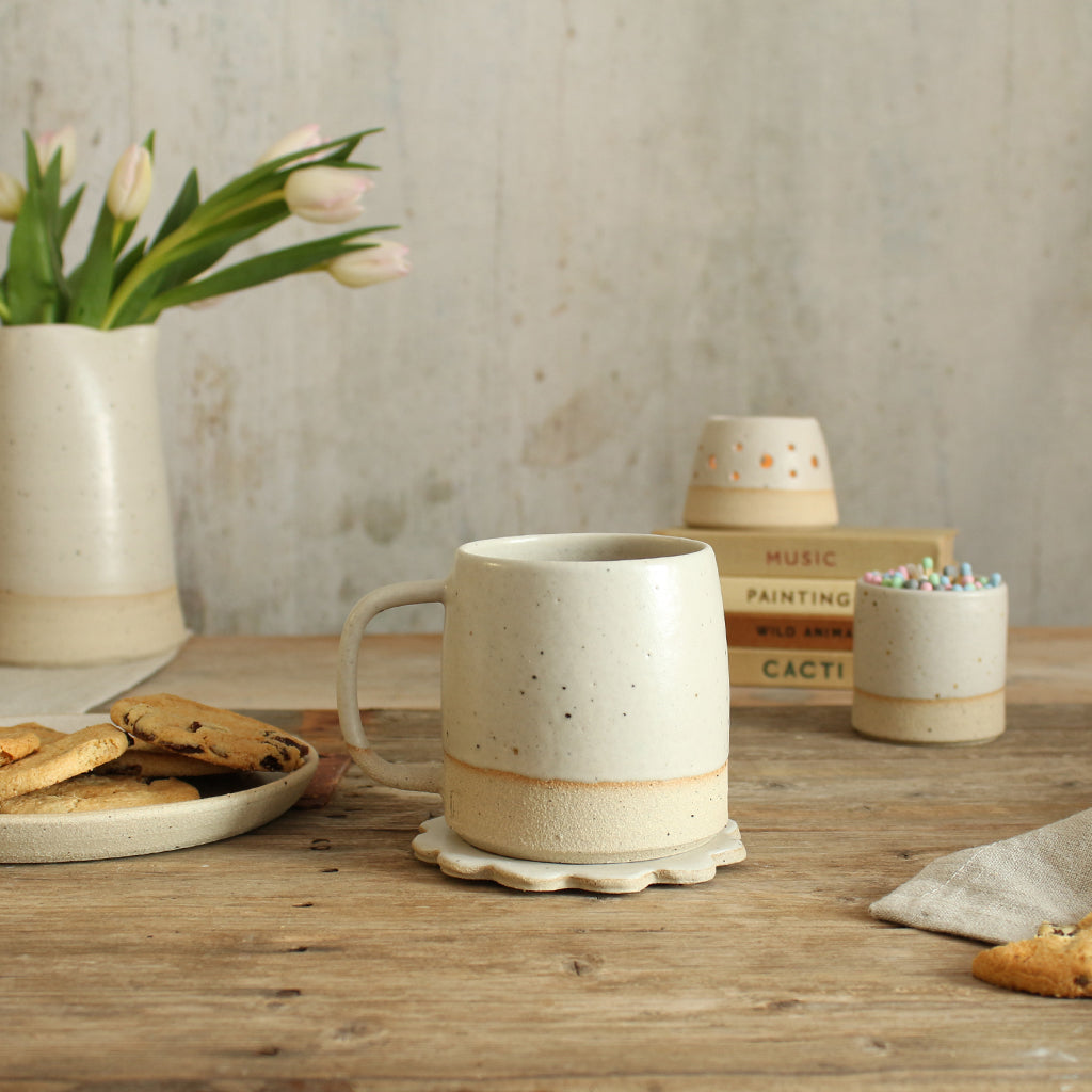 White stoneware mug on white flower coaster with a white match pot, tealight, jug and plate of cookies in background table setting