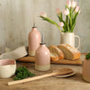 Pink small and large oil pourer on table with bread, wooden spoon, bowls and jug of flowers