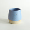 A cornflower blue glazed tumbler with exposed raw flecked clay around the base. Dark flecks from the clay can be seen through the glaze.