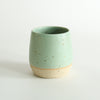 A mint green glazed tumbler with exposed raw flecked clay around the base. Dark flecks from the clay can be seen through the glaze.