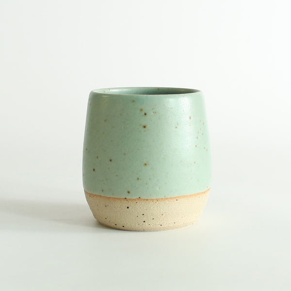 Front view of the mint green glazed tumbler with exposed raw flecked clay around the base. Dark flecks from the clay can be seen through the glaze.