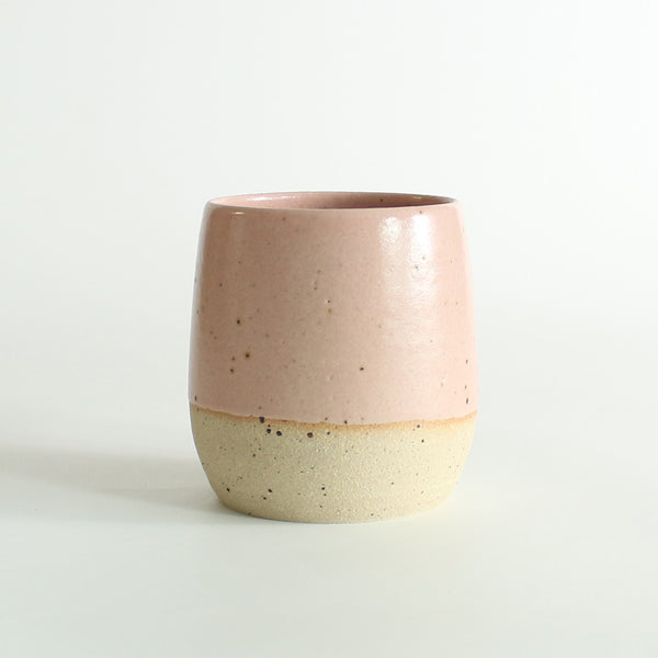 Front view of the pale pink glazed tumbler with exposed raw flecked clay around the base. Dark flecks from the clay can be seen through the glaze.