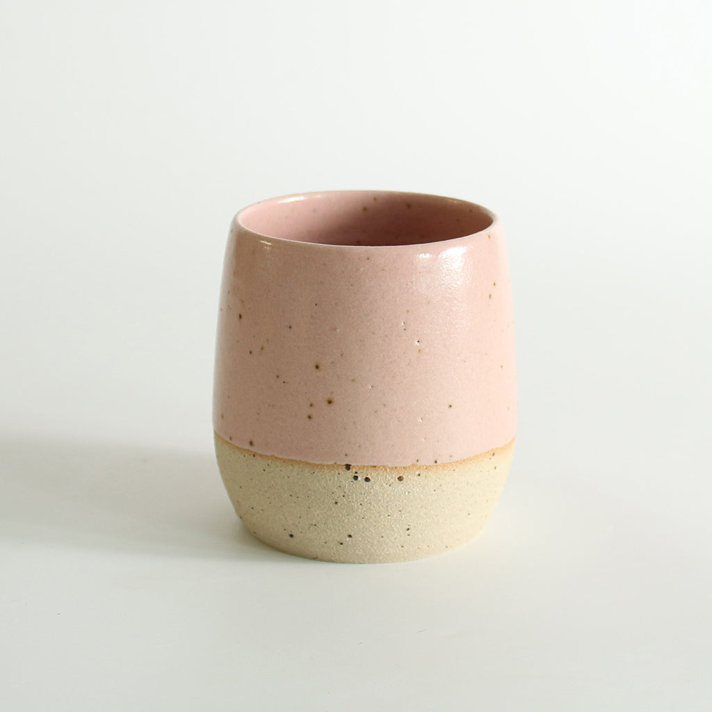 A pale pink glazed tumbler with exposed raw flecked clay around the base. Dark flecks from the clay can be seen through the glaze.