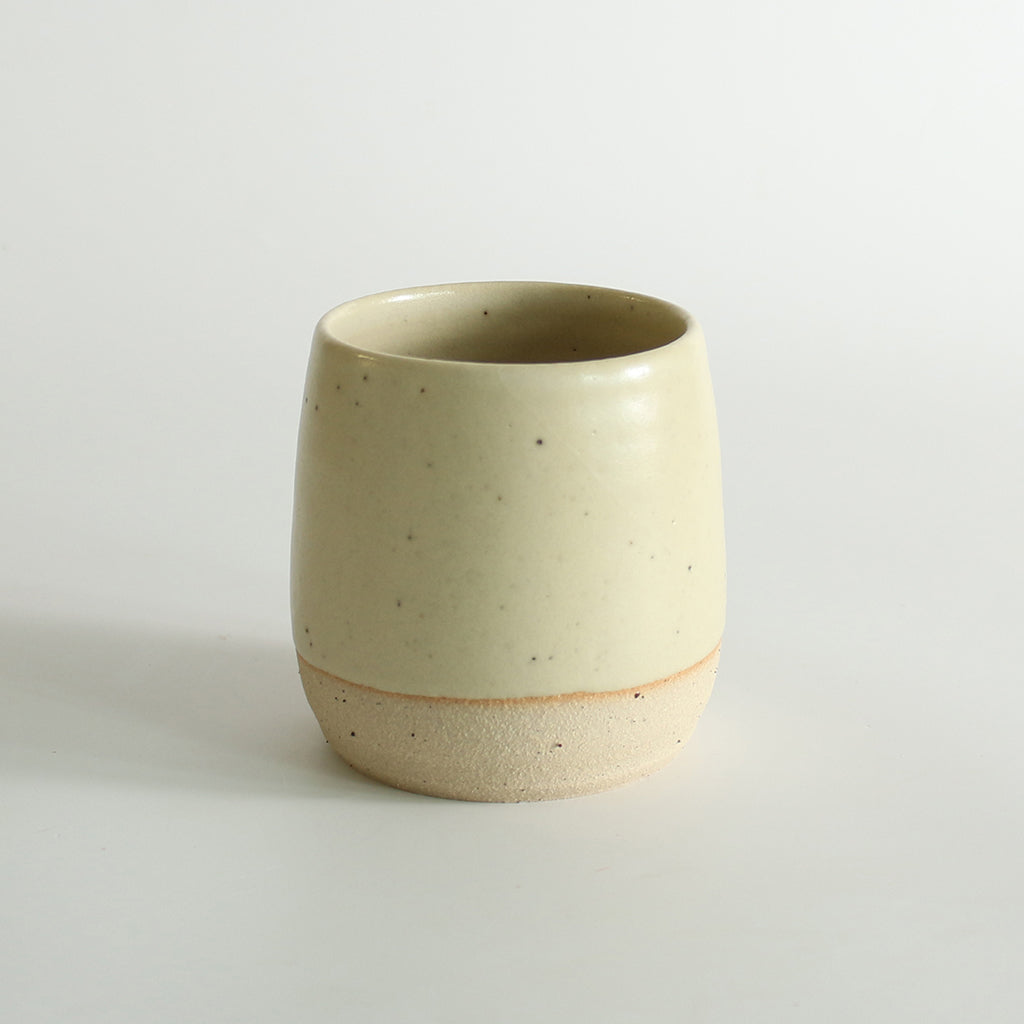 A light yellow glazed tumbler with exposed raw flecked clay around the base. Dark flecks from the clay can be seen through the glaze.