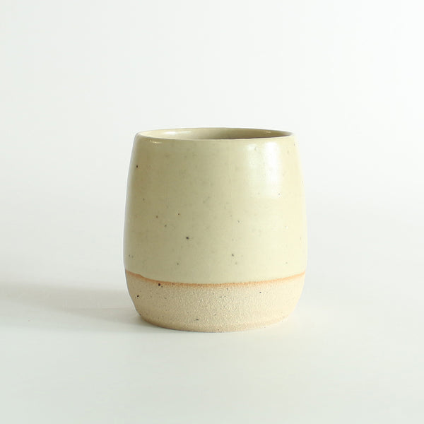 Front view of the light yellow glazed tumbler with exposed raw flecked clay around the base. Dark flecks from the clay can be seen through the glaze.