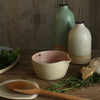 Pink herb bowl on table surrounded by herbs, oil pourers and a spoon on a spoon rest