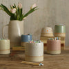 Close up of white match pot in front of other pastel coloured match pots and jug of flowers in background