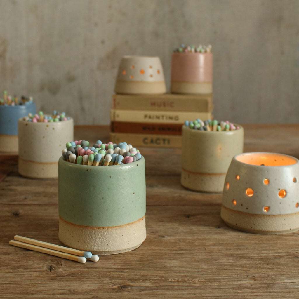 Green match striker pot with pastel matches in front of other pastel pots and lit tealights