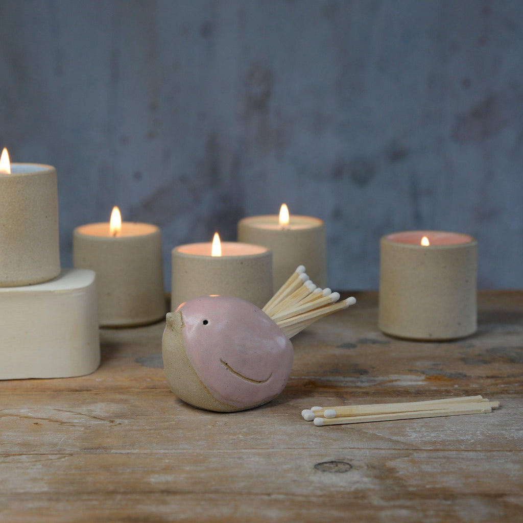 Pink bird pot filled with white matches in front of lit stoneware candles
