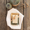 A Moorland soap dish with a natural soap in and dried seed stems. The soap dish is on a linen cloth next to Moorland vases on a dark wooden surface. 